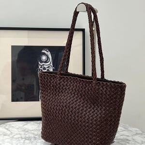 Può includere: Una borsa tote marrone intrecciata con manici intrecciati. La borsa ha una forma rettangolare e una chiusura con cerniera. Il motivo intrecciato è visibile e la borsa sembra essere realizzata in pelle o materiale simile. Anche i manici sono intrecciati.