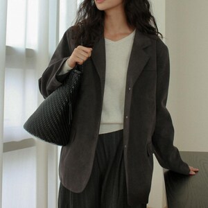 May include: A dark gray blazer with a V-neck sweater and dark pants. The person is holding a black woven handbag with a tassel. The outfit is neutral-toned and suitable for various occasions.