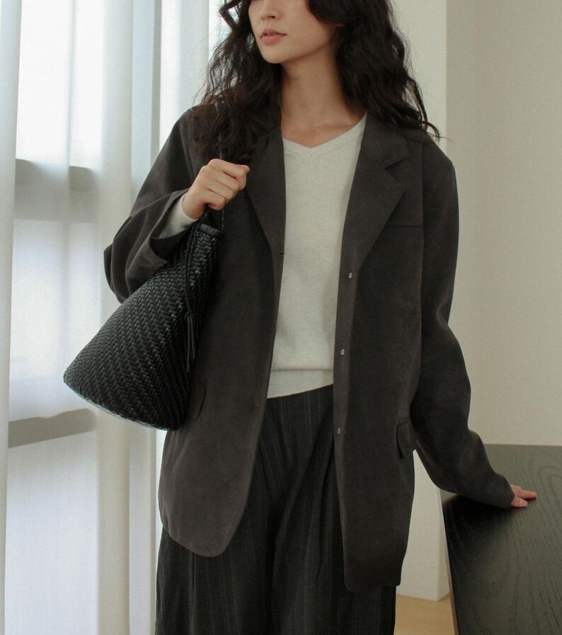 May include: A dark gray blazer with a V-neck sweater and dark pants. The person is holding a black woven handbag with a tassel. The outfit is neutral-toned and suitable for various occasions.