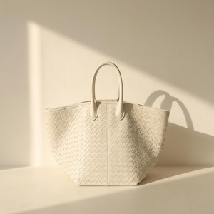 May include: Off-white woven leather tote bag with two top handles. The bag has a trapezoid shape and a textured, basket-weave pattern. The handles are rounded and attached to the top of the bag. The bag is sitting on a white surface.