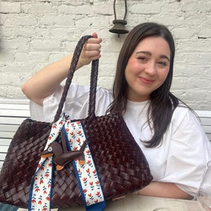 May include: A large, woven, burgundy tote bag with braided handles. A decorative scarf with a red floral pattern and a brown dachshund charm is tied to the handle. The bag is sitting on a table.