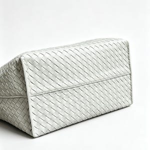 May include: A white, woven leather tote bag. The bag's rectangular shape is visible, with a detailed pattern of interwoven strips creating a textured surface. The bag is set against a plain white background.