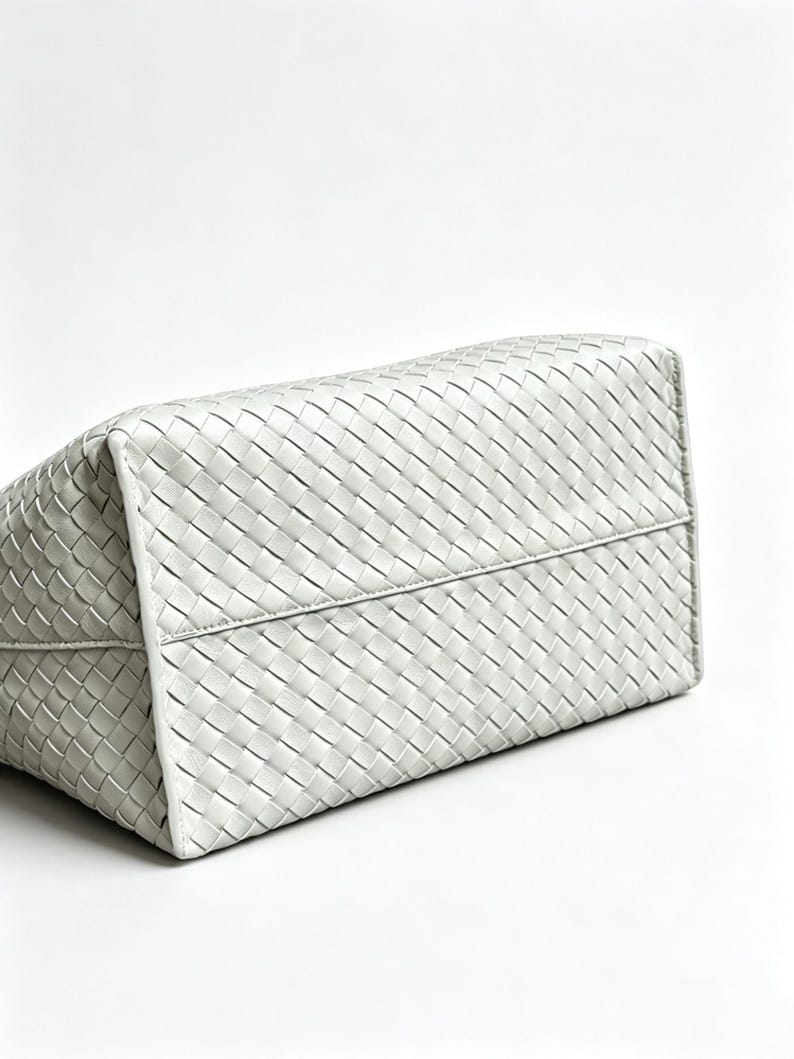 May include: A white, woven leather tote bag. The bag's rectangular shape is visible, with a detailed pattern of interwoven strips creating a textured surface. The bag is set against a plain white background.