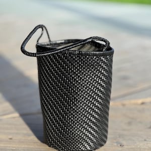 May include: A black woven basket with a handle. The basket is cylindrical and has a textured surface. The handle is arched and attached to the top of the basket. The basket is sitting on a wooden surface.