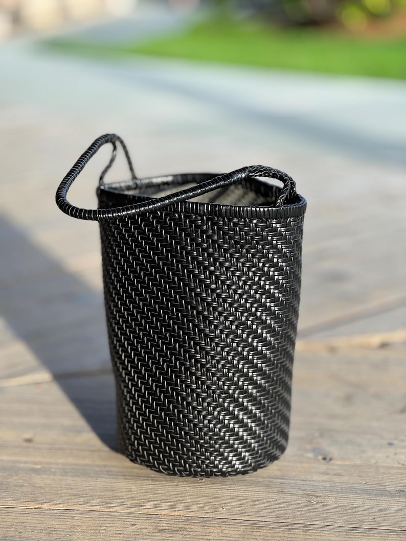 May include: A black woven basket with a handle. The basket is cylindrical and has a textured surface. The handle is arched and attached to the top of the basket. The basket is sitting on a wooden surface.