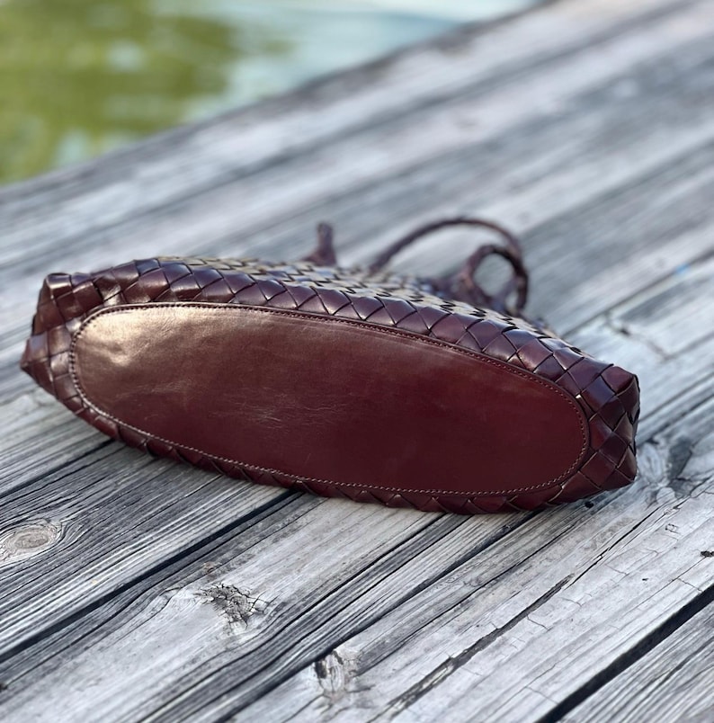 May include: A dark burgundy leather tote bag with a woven design. The bag has a rounded bottom and braided handles. The bag is sitting on a wooden surface.