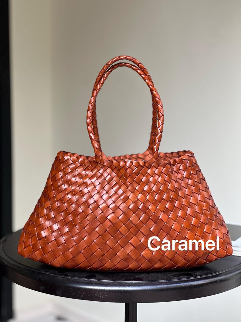 May include: A caramel-colored woven tote bag with braided handles. The bag is made of a tightly woven material, creating a textured appearance. The word "Caramel" is printed on the bag.