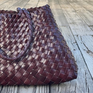 May include: A burgundy-colored woven leather tote bag. The bag features a basketweave design and a braided handle. The bag is sitting on a weathered wooden surface.