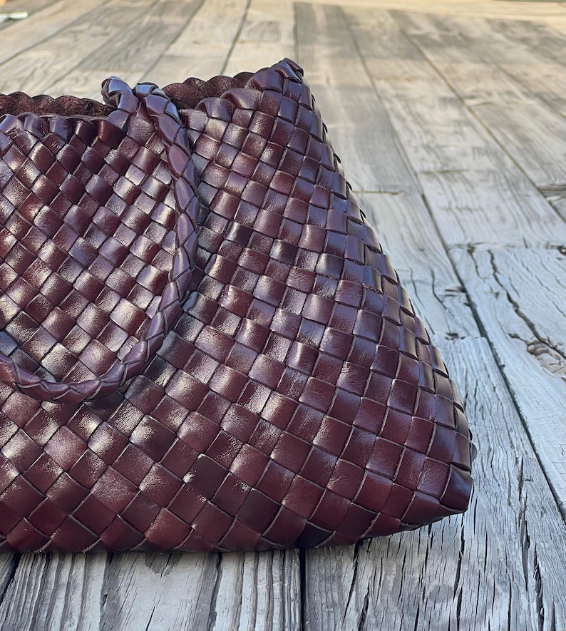 May include: A burgundy-colored woven leather tote bag. The bag features a basketweave design and a braided handle. The bag is sitting on a weathered wooden surface.