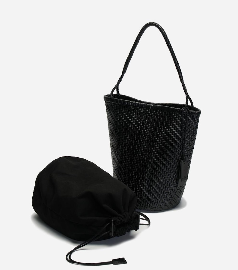 May include: A black woven bucket bag with a matching drawstring pouch. The bag has a single handle and a small tag. The weave pattern is visible on the bag's exterior. The pouch is made of a soft, opaque material.