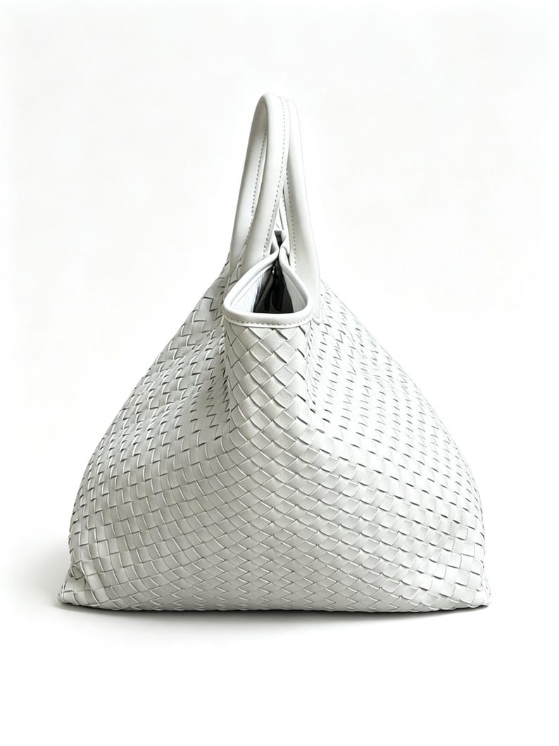 May include: A white woven leather tote bag with a single handle. The bag has a large, open top and a woven pattern throughout the body. The handle is curved and attached to the top of the bag. The bag is made of a soft, pliable leather.