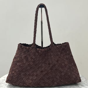 Peut inclure: Un sac fourre-tout marron tressé avec des anses tressées. Le sac a une forme trapézoïdale et une large ouverture. Le matériau semble être du daim ou un tissu doux similaire. Le sac est posé sur une table en marbre.