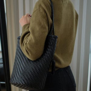 May include: A black woven tote bag with a shoulder strap. The bag has a rectangular shape and a textured, basket-weave design. The bag is being held by a person wearing a green sweater and black jeans.