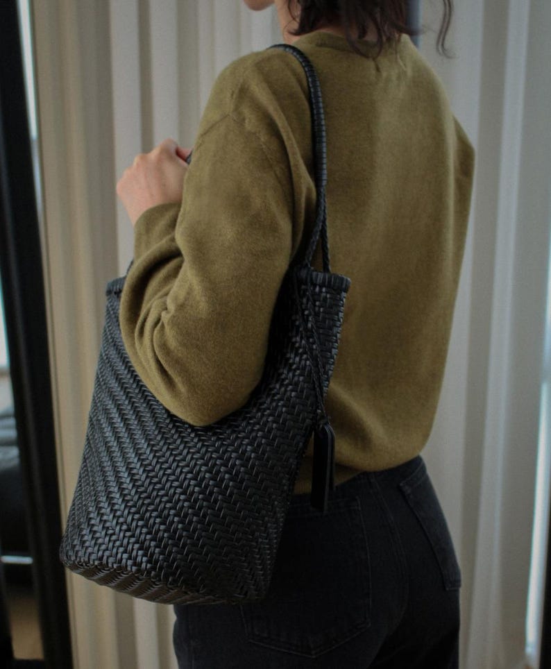 May include: A black woven tote bag with a shoulder strap. The bag has a rectangular shape and a textured, basket-weave design. The bag is being held by a person wearing a green sweater and black jeans.