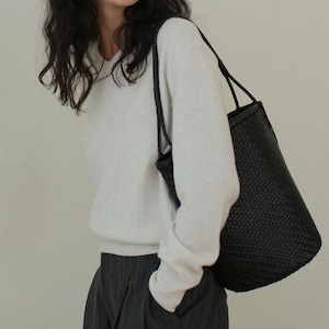 May include: A black woven tote bag with long handles. The bag is carried over the shoulder. The bag has a cylindrical shape and a textured surface. The person is wearing a white sweater and gray pants.