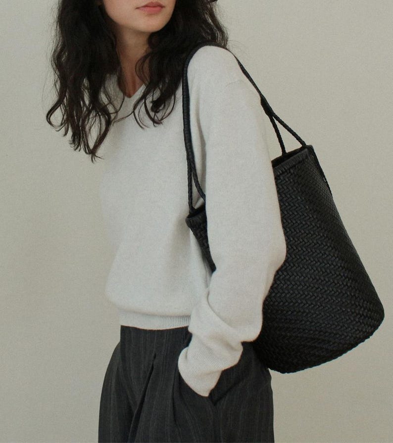 May include: A black woven tote bag with long handles. The bag is carried over the shoulder. The bag has a cylindrical shape and a textured surface. The person is wearing a white sweater and gray pants.