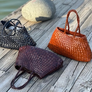 May include: Three woven leather tote bags in black, reddish-brown, and orange. The bags have braided handles and a basket-weave design. The bags are displayed on a wooden surface, with a large gray stone in the background.