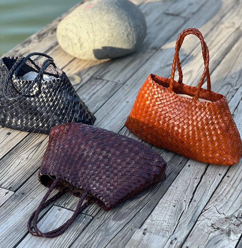 May include: Three woven leather tote bags in black, reddish-brown, and orange. The bags have braided handles and a basket-weave design. The bags are displayed on a wooden surface, with a large gray stone in the background.