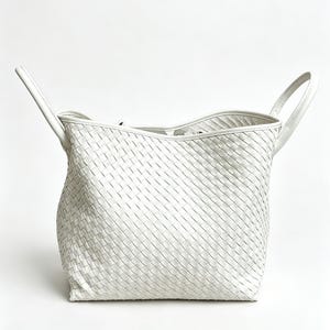 May include: A white woven leather tote bag with two top handles. The bag has a rectangular shape with a woven pattern throughout the body. The handles are attached to the top of the bag and are curved.