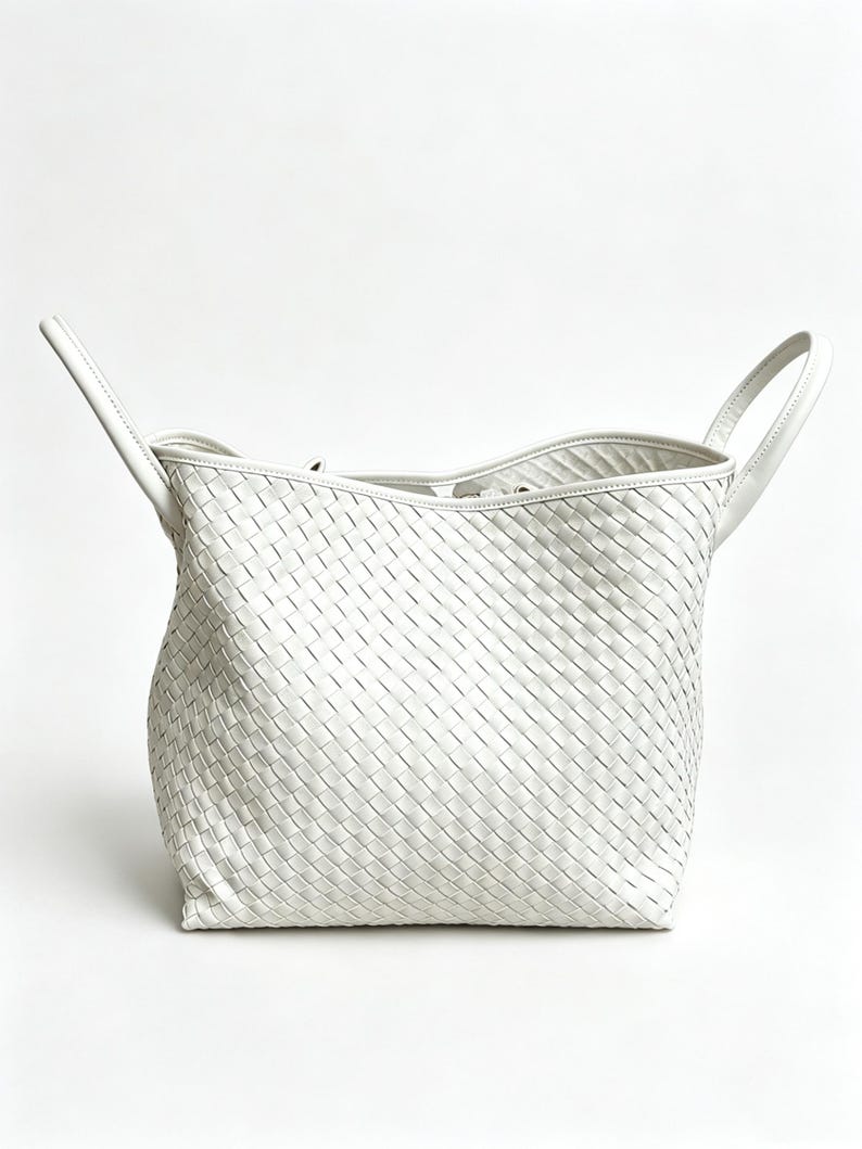 May include: A white woven leather tote bag with two top handles. The bag has a rectangular shape with a woven pattern throughout the body. The handles are attached to the top of the bag and are curved.