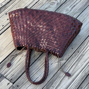 May include: A burgundy-colored woven leather tote bag with braided handles. The bag has a rectangular shape with a wide opening and a drawstring closure. The bag is sitting on a wooden surface.