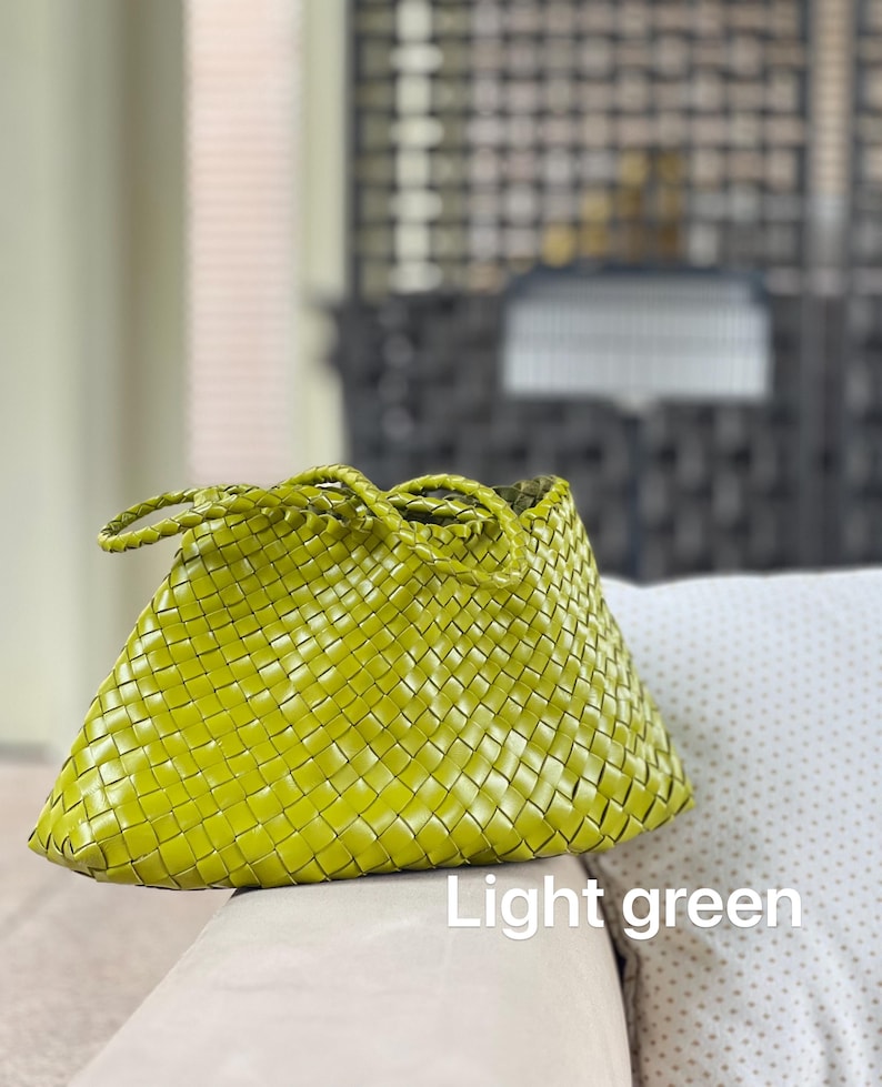 May include: Light green woven handbag with braided handles. The bag features a basket weave design and a trapezoid shape. The text "Light green" is visible at the bottom of the image. The bag is sitting on a light-colored surface.