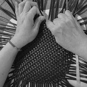 May include: Black and white image of hands weaving a basket. The hands are working with dark, thin strips, interlacing them to form a woven pattern. The basket's base is visible, with the weaving process in progress.