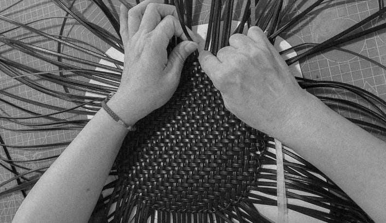 May include: Black and white image of hands weaving a basket. The hands are working with dark, thin strips, interlacing them to form a woven pattern. The basket's base is visible, with the weaving process in progress.