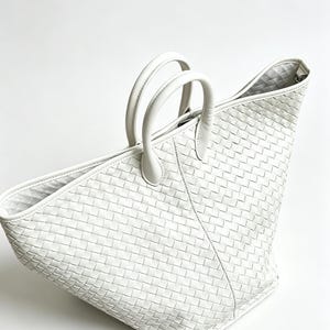 May include: A white woven leather tote bag with two rounded handles. The bag has a trapezoid shape and a textured, basket-weave pattern. The interior lining is visible at the top. The bag is set against a white background.