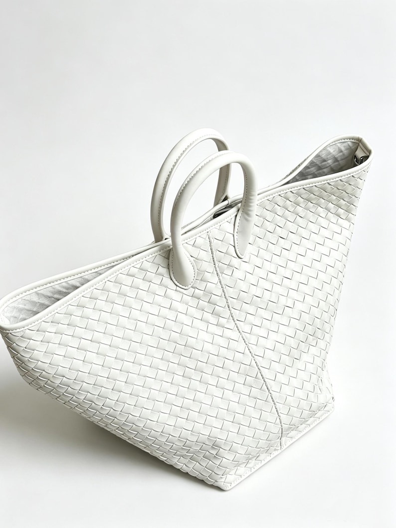 May include: A white woven leather tote bag with two rounded handles. The bag has a trapezoid shape and a textured, basket-weave pattern. The interior lining is visible at the top. The bag is set against a white background.