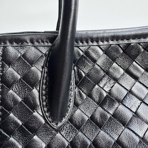 May include: Close-up of a black leather handbag with a woven pattern. The bag features a sturdy handle and a detailed, textured surface. The stitching is visible along the edges, highlighting the craftsmanship of the bag.
