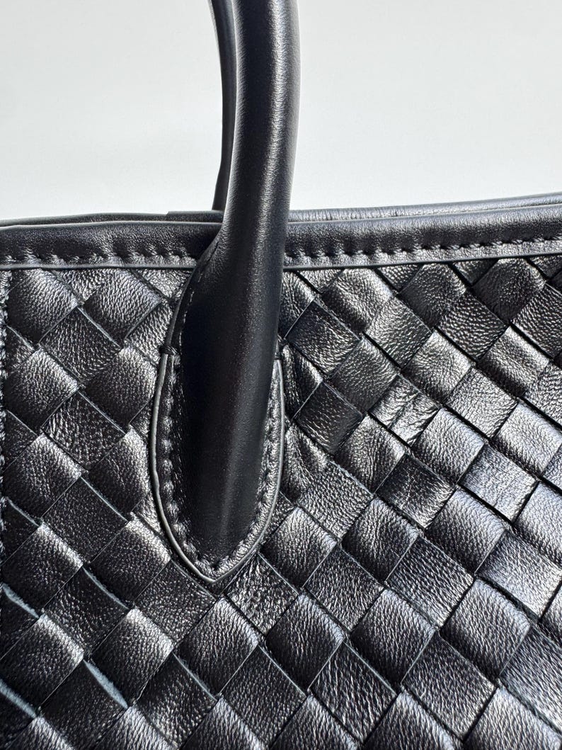 May include: Close-up of a black leather handbag with a woven pattern. The bag features a sturdy handle and a detailed, textured surface. The stitching is visible along the edges, highlighting the craftsmanship of the bag.