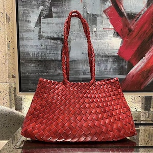 Puede incluir: Un bolso tote de color rojo intenso que presenta un intrincado patrón de cuero trenzado en todo el cuerpo. El bolso incluye dos correas de hombro de cuero trenzado a juego que se extienden desde el borde superior, creando una apariencia texturizada uniforme.
