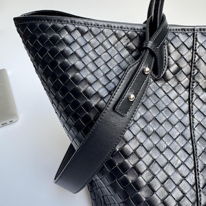 May include: Black leather tote bag with a woven pattern. The bag features a shoulder strap and two top handles. The bag is made of a woven leather material and has a structured shape. The bag is a dark black color.