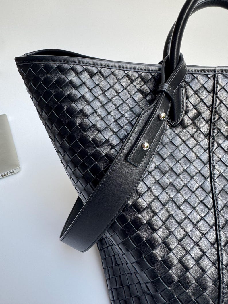 May include: Black leather tote bag with a woven pattern. The bag features a shoulder strap and two top handles. The bag is made of a woven leather material and has a structured shape. The bag is a dark black color.