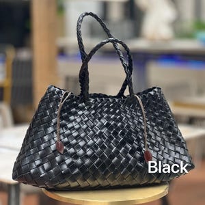 May include: Black woven tote bag with braided handles and brown leather accents. The bag features a geometric pattern and a rectangular shape. The word "Black" is printed on the bag.