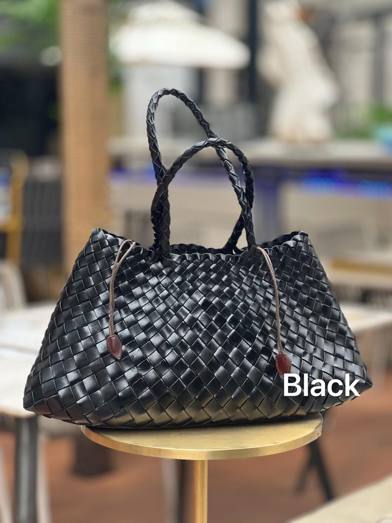 May include: Black woven tote bag with braided handles and brown leather accents. The bag features a geometric pattern and a rectangular shape. The word "Black" is printed on the bag.