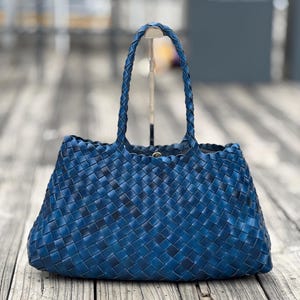 May include: A deep blue tote bag featuring a woven basket-weave pattern throughout the body and two braided handles. The bag has a wide, rectangular base and a slightly tapered top opening with a visible metallic snap closure at the center.