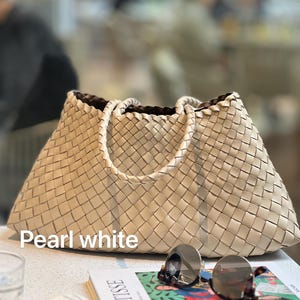May include: Pearl white woven tote bag with braided handles. The bag is rectangular with a trapezoid shape. The bag is sitting on a white surface next to a pair of sunglasses and a book with the word "MATISSE" on the cover.