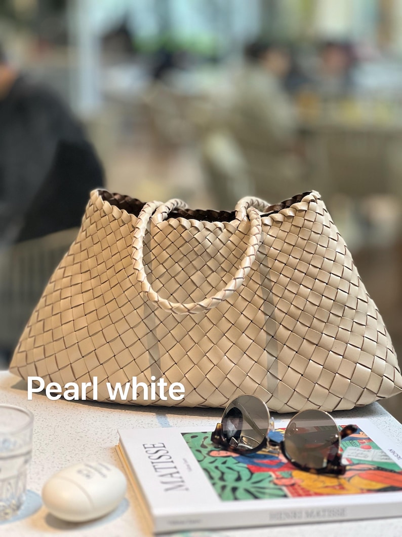 May include: Pearl white woven tote bag with braided handles. The bag is rectangular with a trapezoid shape. The bag is sitting on a white surface next to a pair of sunglasses and a book with the word "MATISSE" on the cover.