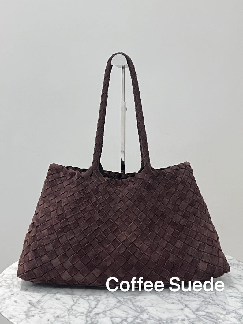 May include: A brown suede tote bag with a woven design. The bag has two braided handles and the text "Coffee Suede" at the bottom. The bag is displayed on a white surface.
