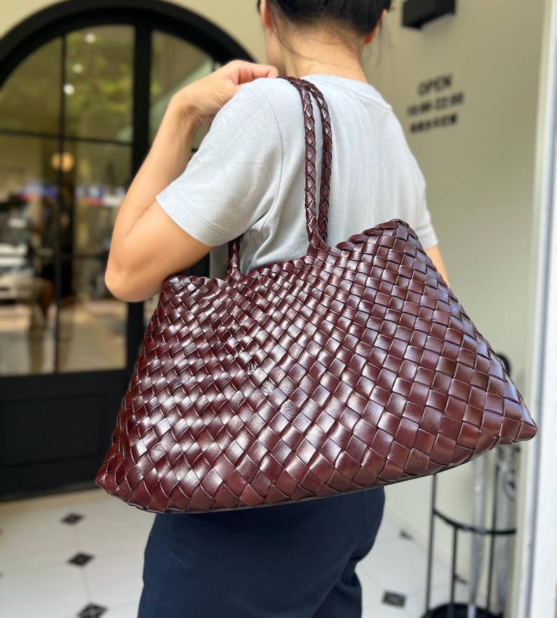 May include: A large, woven leather tote bag in a rich burgundy color. The bag features a braided handle and a spacious interior, perfect for carrying everyday essentials. The weave pattern creates a textured surface.
