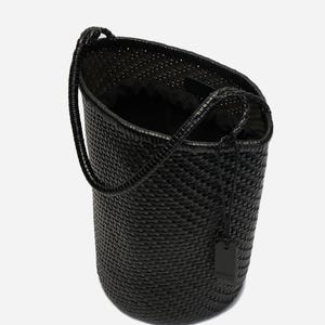 May include: A black woven tote bag with a cylindrical shape and a single handle. The bag has a textured surface and a small tag hanging from the handle. The interior of the bag is black.