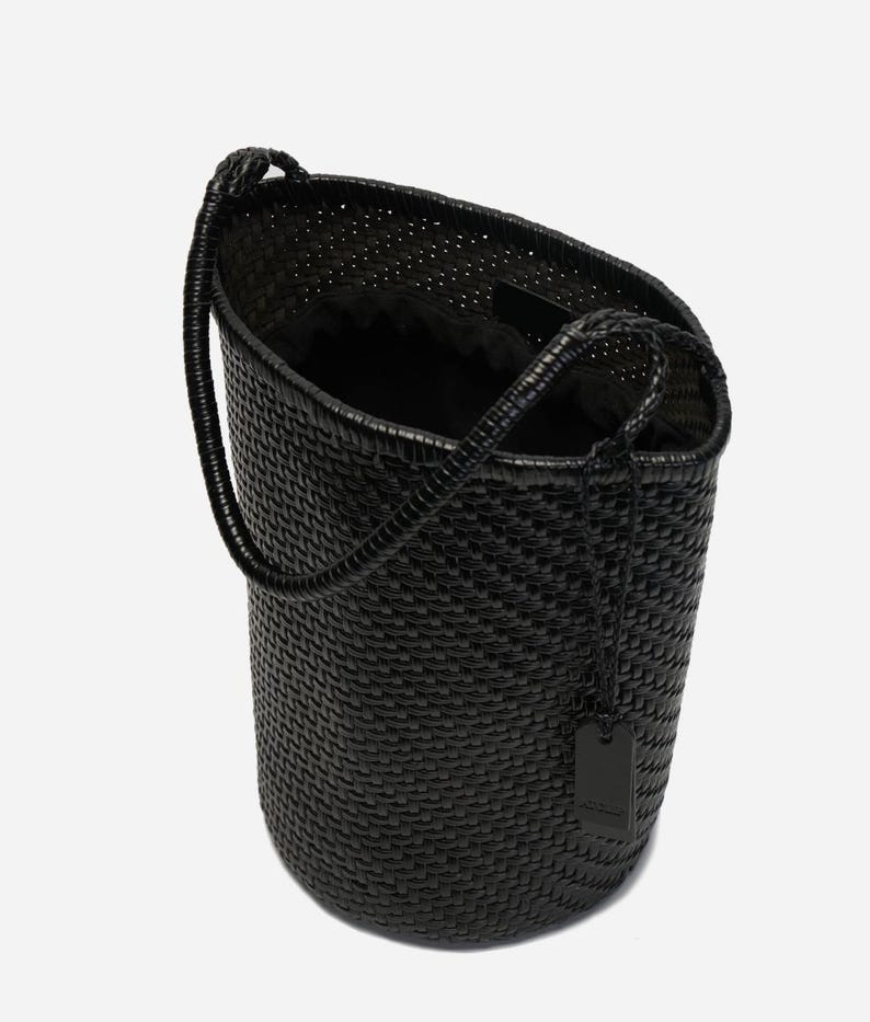May include: A black woven tote bag with a cylindrical shape and a single handle. The bag has a textured surface and a small tag hanging from the handle. The interior of the bag is black.