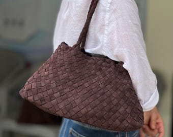 Può includere: Una borsa a tracolla in pelle intrecciata marrone con una tracolla intrecciata. La borsa ha una consistenza morbida, simile alla pelle scamosciata, e una forma trapezoidale. La borsa è portata a spalla.