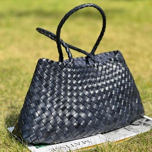 May include: A black woven leather tote bag with a trapezoid shape and braided handles. The bag is sitting on a white surface with text and green grass in the background. The bag has a structured design.