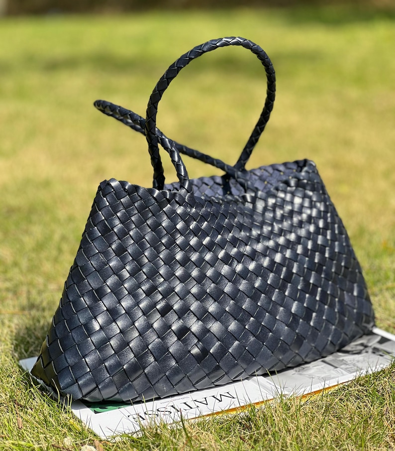 May include: A black woven leather tote bag with a trapezoid shape and braided handles. The bag is sitting on a white surface with text and green grass in the background. The bag has a structured design.