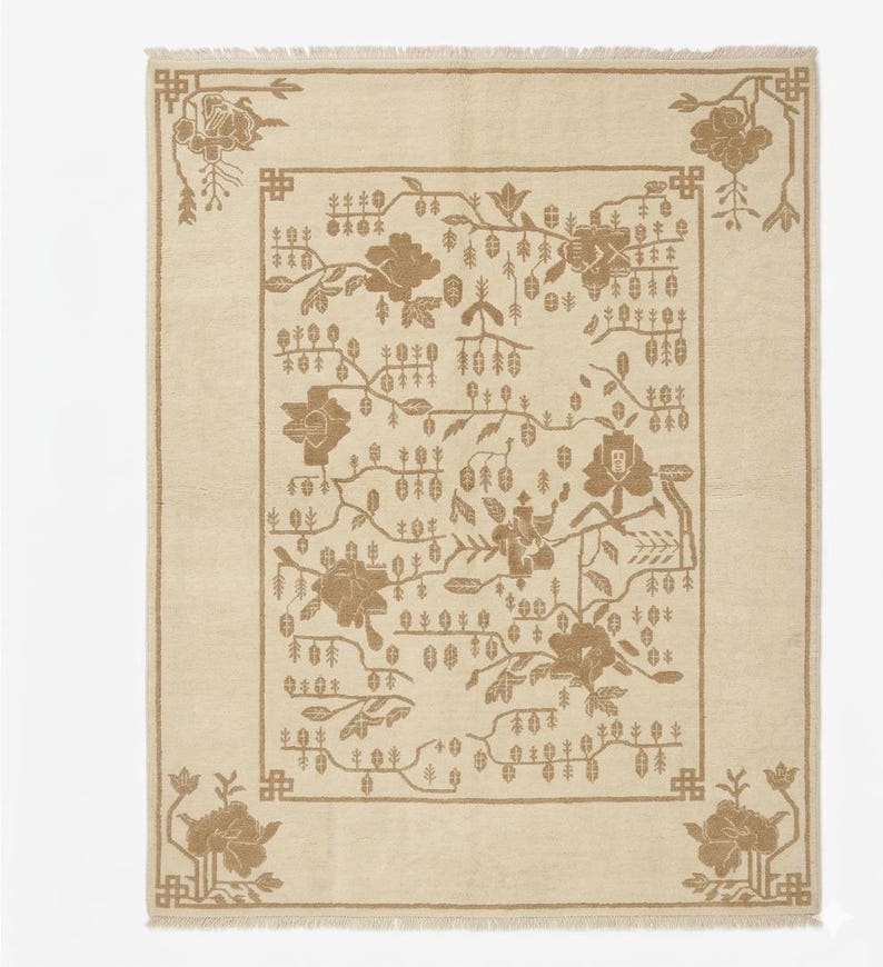 May include: Cream-coloured rectangular rug with a floral pattern in brown tones. The rug has a border and corner details with stylised flowers and foliage. The central design features flowers, branches, and small leaf-like shapes.