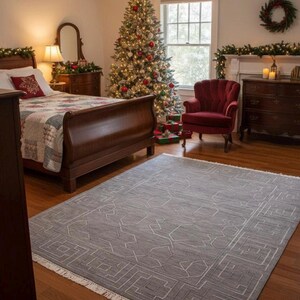 Puede incluir: Un acogedor dormitorio con un árbol de Navidad decorado, un sillón rojo y una alfombra gris con diseño geométrico. La habitación cuenta con una cama con colcha, una cómoda y una ventana con una corona. El árbol está adornado con luces y adornos.