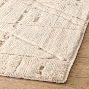 May include: Cream-colored, textured rug with a geometric pattern. The rug features a series of raised, rectangular and triangular shapes, creating a tactile surface. The edges are neatly finished with a subtle border. Ideal for adding warmth and texture to any room.